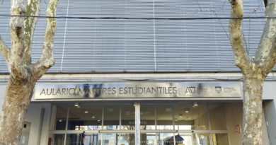 Fachada del Aulario de Mártires Estudiantiles, ubicado en Gonzalo Ramírez 1915. Se observa la entrada principal del edificio con puertas de vidrio, cartelería institucional en la parte superior y logos de la Universidad de la República. En primer plano hay árboles a ambos lados de la vereda.