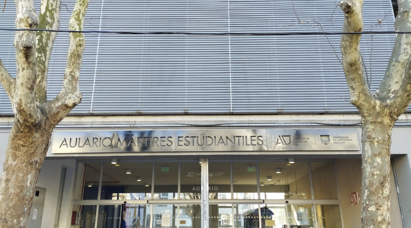 Fachada del Aulario de Mártires Estudiantiles, ubicado en Gonzalo Ramírez 1915. Se observa la entrada principal del edificio con puertas de vidrio, cartelería institucional en la parte superior y logos de la Universidad de la República. En primer plano hay árboles a ambos lados de la vereda.