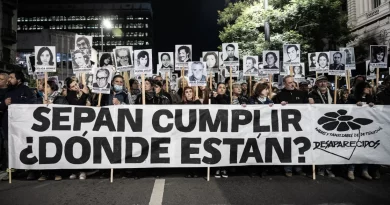 Fotografía nocturna de la marcha del 20 de Silencio, multitudinaria sobre la avenida 18 de julio. En primer plano, un gran pasacalle blanco ocupa casi todo el ancho de la imagen, con letras negras enormes que dicen: “SEPAN CUMPLIR ¿DÓNDE ESTÁN?”. A la derecha del cartel aparece el logo de Madres y Familiares de Detenidos Desaparecidos, compuesto por una flor negra y el texto “Desaparecidos”. Detrás de la pancarta hay decenas de personas caminando juntas. Muchas sostienen retratos en blanco y negro de personas desaparecidas, montados sobre palos altos. En cada retrato se leen nombres impresos debajo de las fotografías. Las expresiones son serias y concentradas. Algunas personas usan abrigos, bufandas y gorros, lo que sugiere una noche fría.