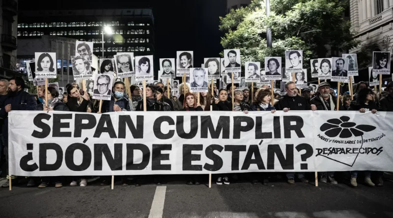 Fotografía nocturna de la marcha del 20 de Silencio, multitudinaria sobre la avenida 18 de julio. En primer plano, un gran pasacalle blanco ocupa casi todo el ancho de la imagen, con letras negras enormes que dicen: “SEPAN CUMPLIR ¿DÓNDE ESTÁN?”. A la derecha del cartel aparece el logo de Madres y Familiares de Detenidos Desaparecidos, compuesto por una flor negra y el texto “Desaparecidos”. Detrás de la pancarta hay decenas de personas caminando juntas. Muchas sostienen retratos en blanco y negro de personas desaparecidas, montados sobre palos altos. En cada retrato se leen nombres impresos debajo de las fotografías. Las expresiones son serias y concentradas. Algunas personas usan abrigos, bufandas y gorros, lo que sugiere una noche fría.